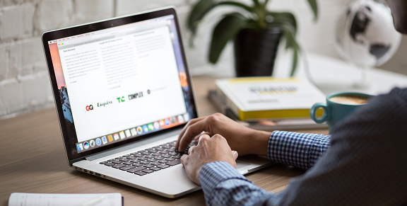 Employee working at a laptop on a wooden desk against a white washed wall with books, a plant and a hot beverage