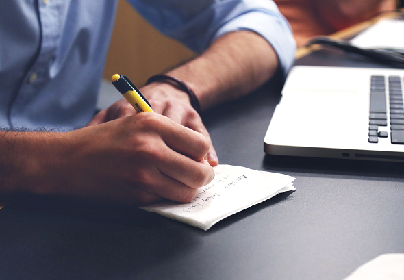 Employee writes list on paper with pen next to open laptop