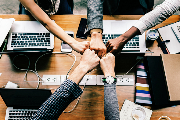 Employees touch fists above a wooden table with open laptops placed upon it