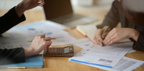 A colleague enters figures into a calculator over a desk strewn with financial reports.