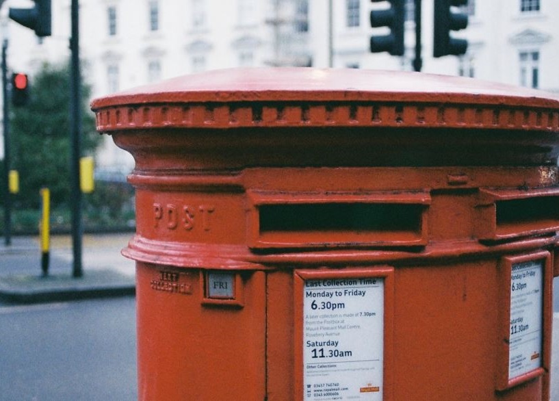 Top section of a red post box on a street with traffic lights behind