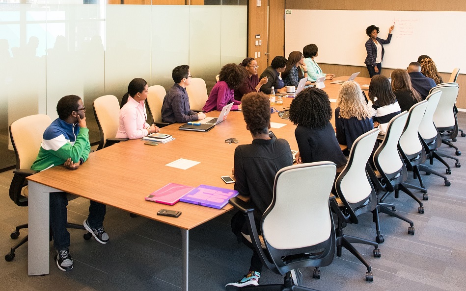 A busy meeting room with attendees facing a presenter against a blank white board