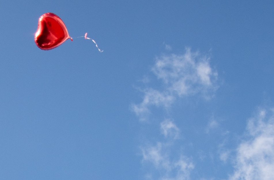 Foil heart balloon floating upwards in the top left against a blue sky with a whisp of white cloud stemming from the opposite corner